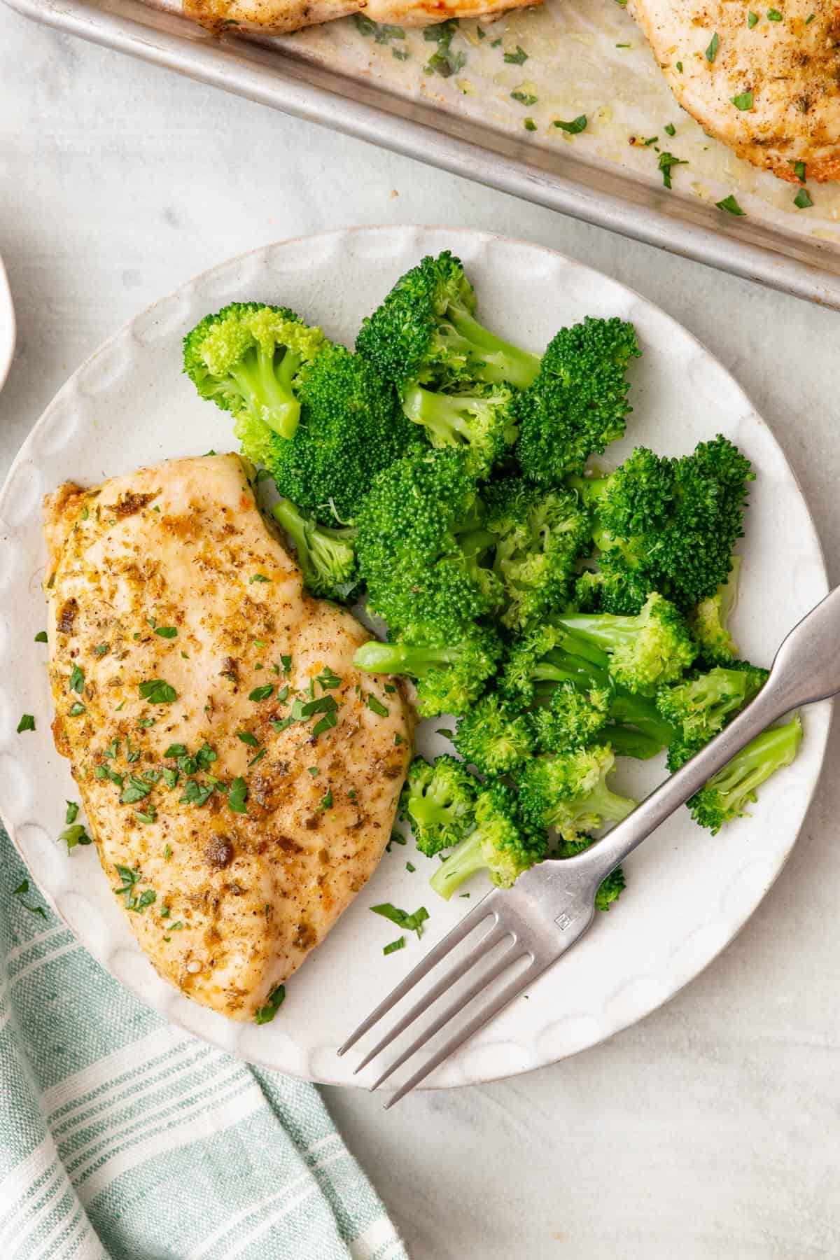 A white plate with baked ranch chicken and a side of broccoli with a tray of other baked chicken and a seafoam green and white striped cloth napkin nearby.