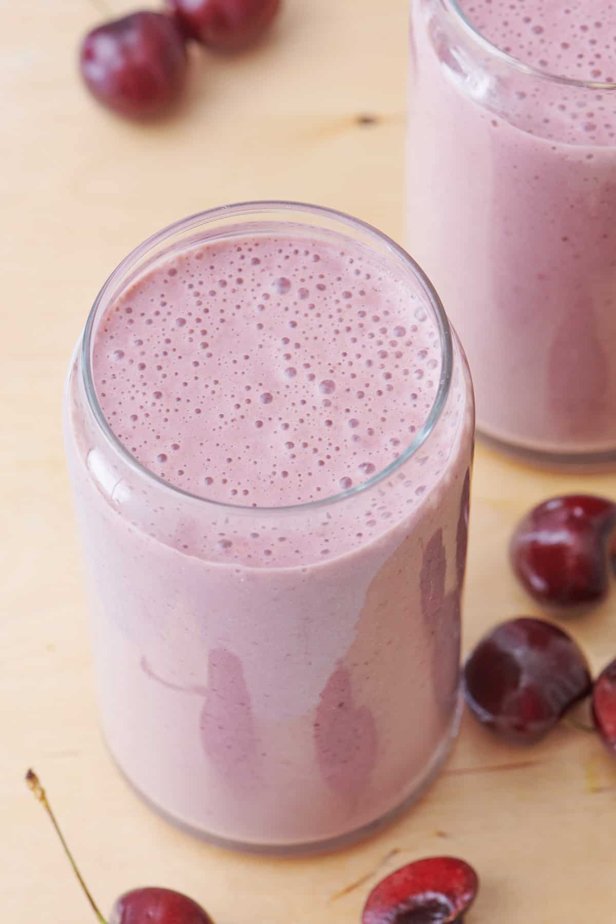 Overhead shot of cherry smoothie in a glass, surrounded by cherries with another glass of smoothie nearby.