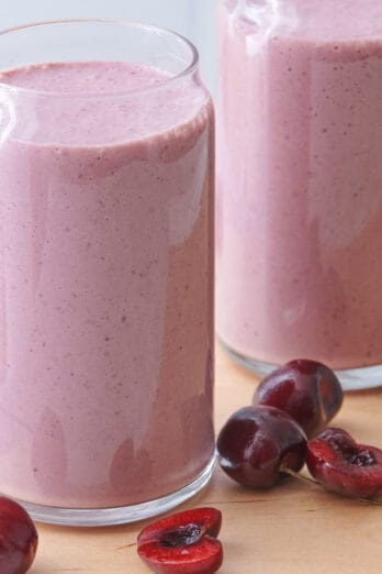 2 glasses of cherry smoothie with cherries surrounding them.
