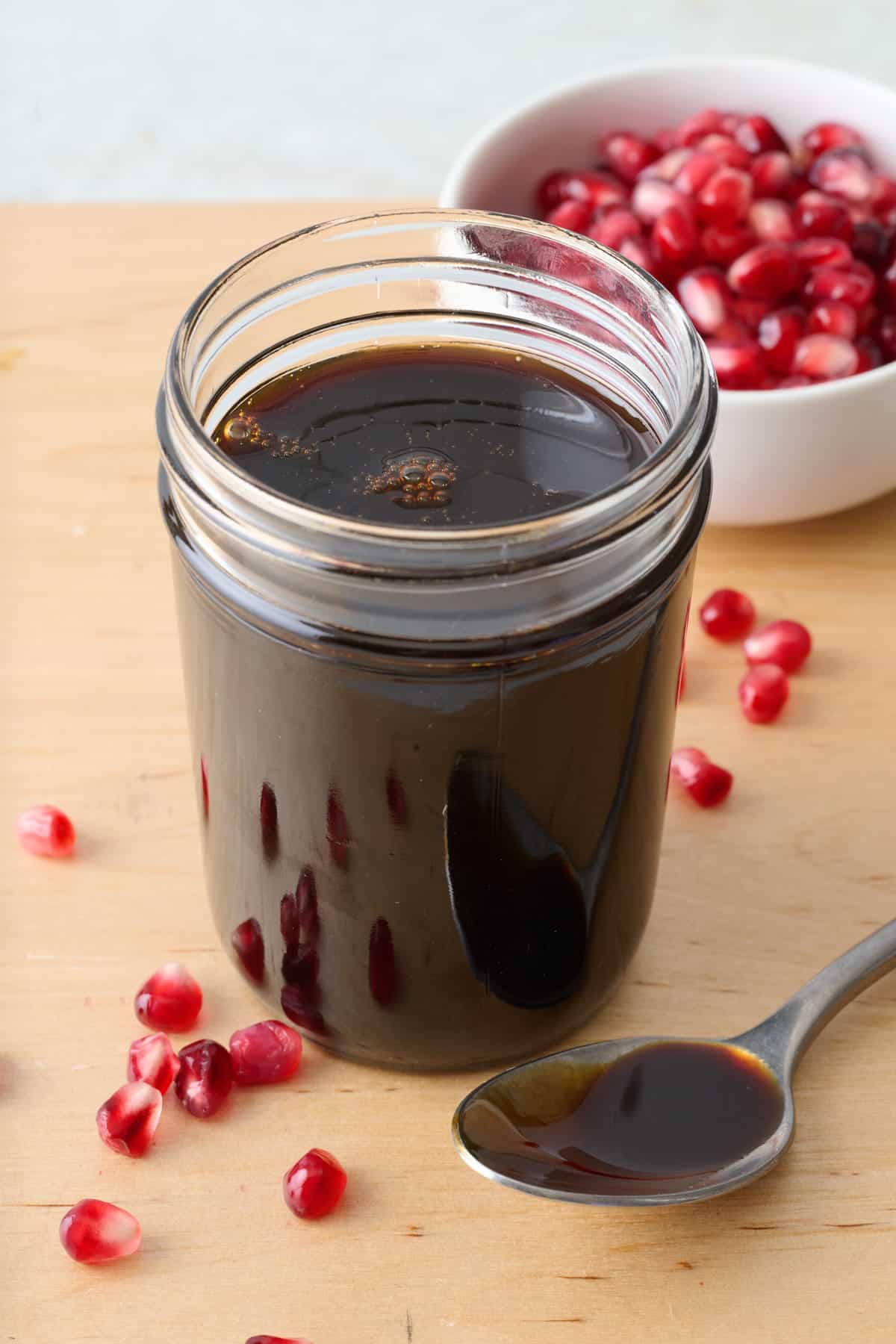 Small jar of thick homemade pomegranate molasses with a spoonful nearby and extra pomegranate seeds in a bowl.