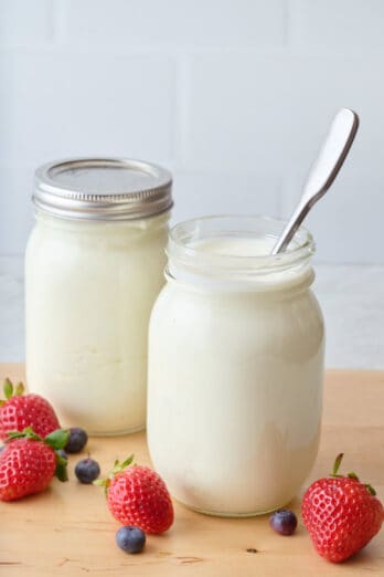 Strawberries and blueberries surrounding 2 mason jars of Instant Pot yogurt: 1 covered with a lid, 1 with a spoon in it.