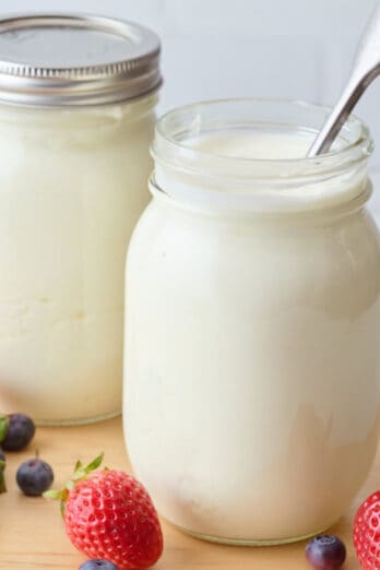 Strawberries and blueberries surrounding 2 mason jars of Instant Pot yogurt: 1 with a lid, 1 with a spoon.