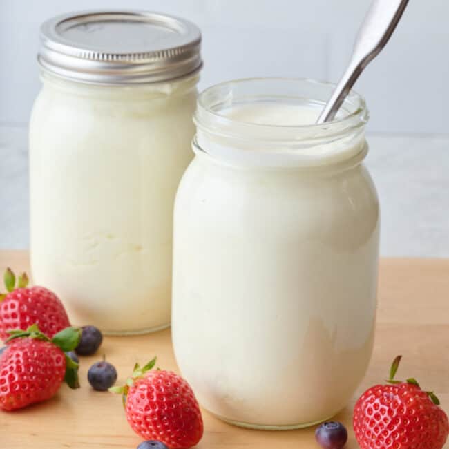 Strawberries and blueberries surrounding 2 mason jars of Instant Pot yogurt: 1 with a lid, 1 with a spoon.
