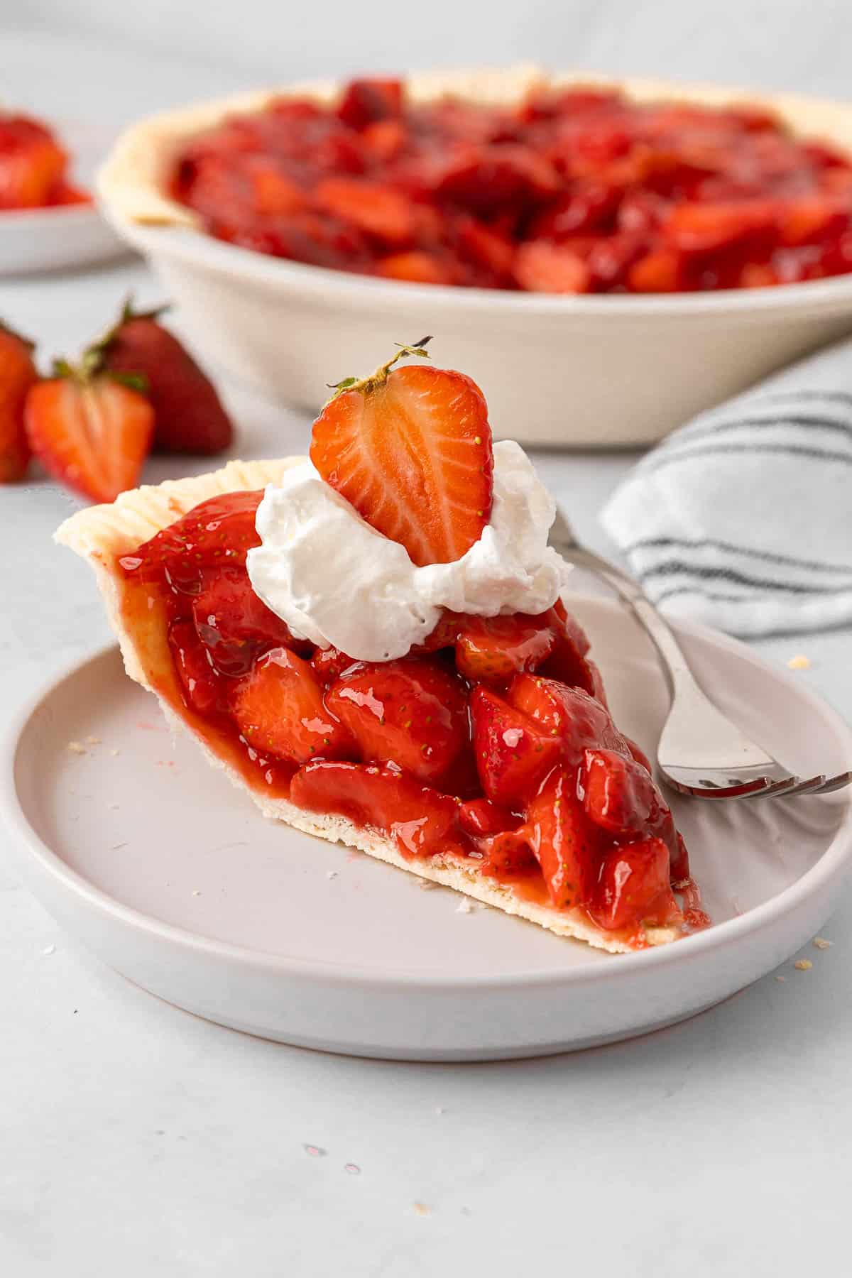 A slice of strawberry pie with whipped cream and a fresh strawberry on top on a small plate.