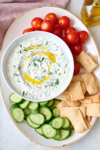 Tzatziki sauce on a platter surrounded by cherry tomatoes, sliced Persian cucumbers, and pita chips.