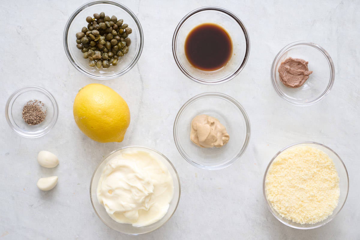 Small bowls of ingredients for Caesar salad dressing, including grated cheese, mayonnaise, and mustard.