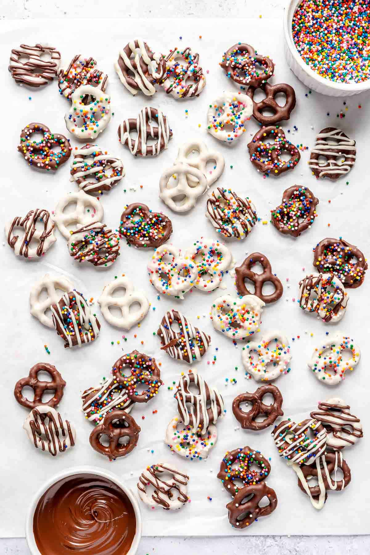 Chocolate-covered pretzels on parchment paper with bowl of chocolate and bowl of rainbow sprinkles nearby.