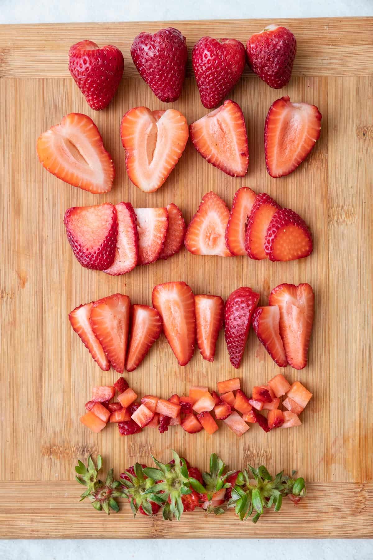 How to cut strawberries 4 ways: Whole strawberries, halved strawberries, sliced strawberries, quartered strawberries, diced strawberries, and green strawberry tops on a wooden cutting board.