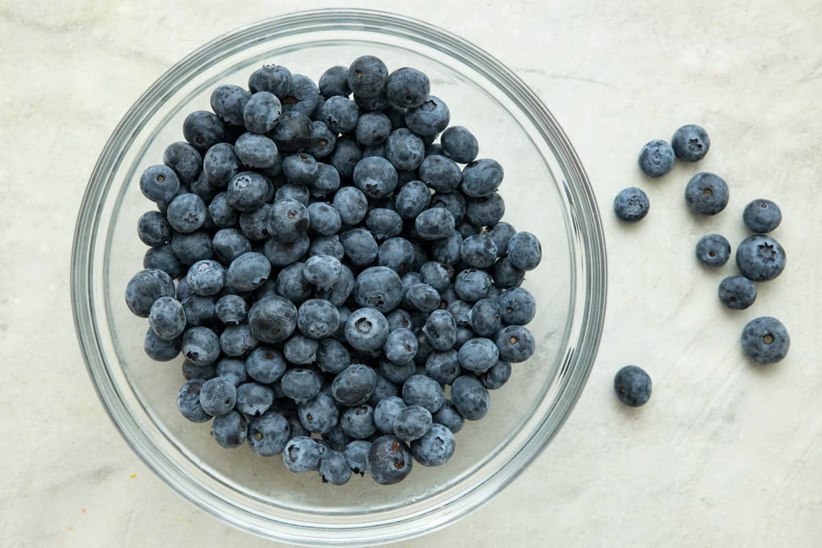 Glass mixing bowl of fresh blueberries on marble countertop.
