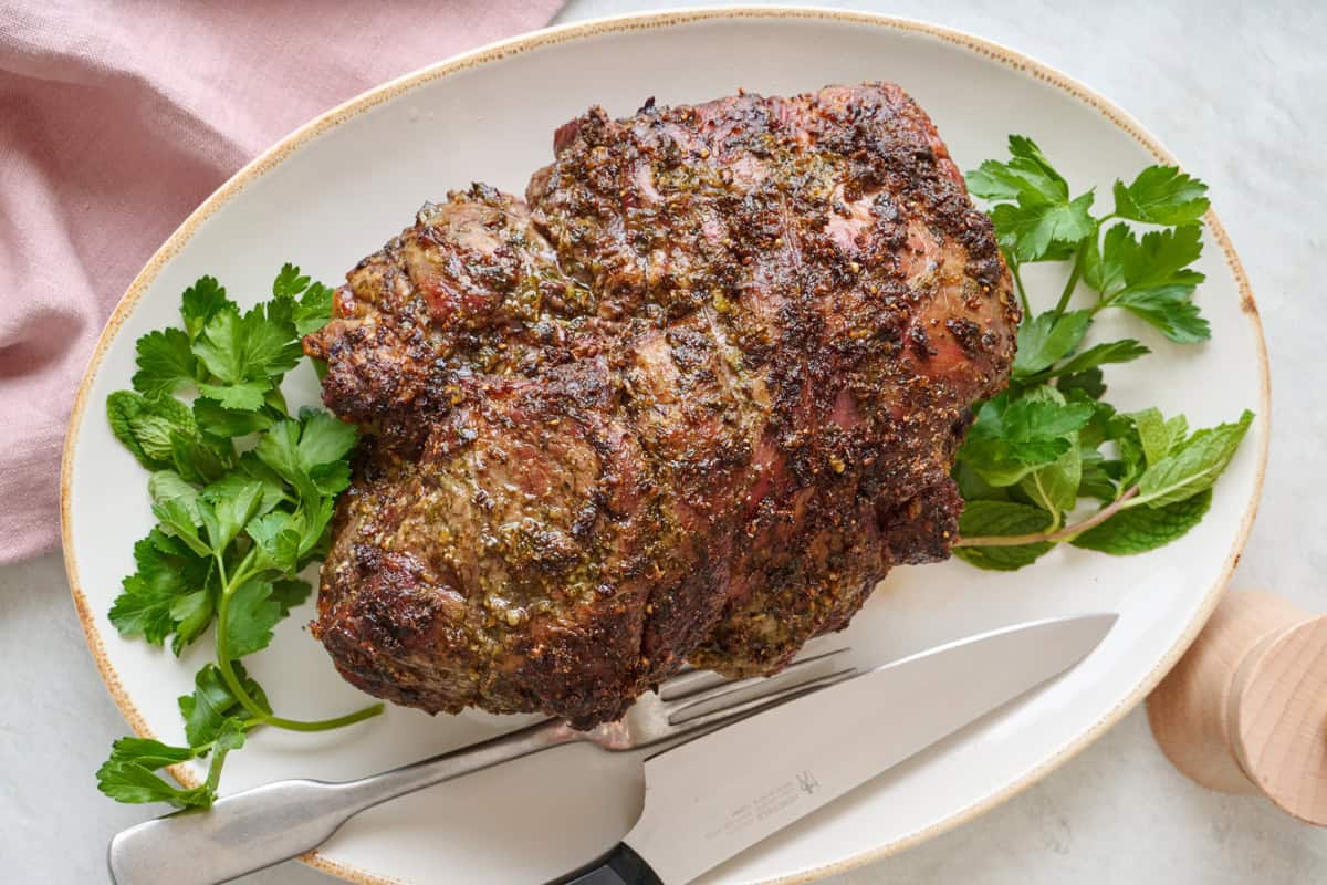 Roasted leg of lamb on a platter with fresh parsley, a fork, and a carving knife.