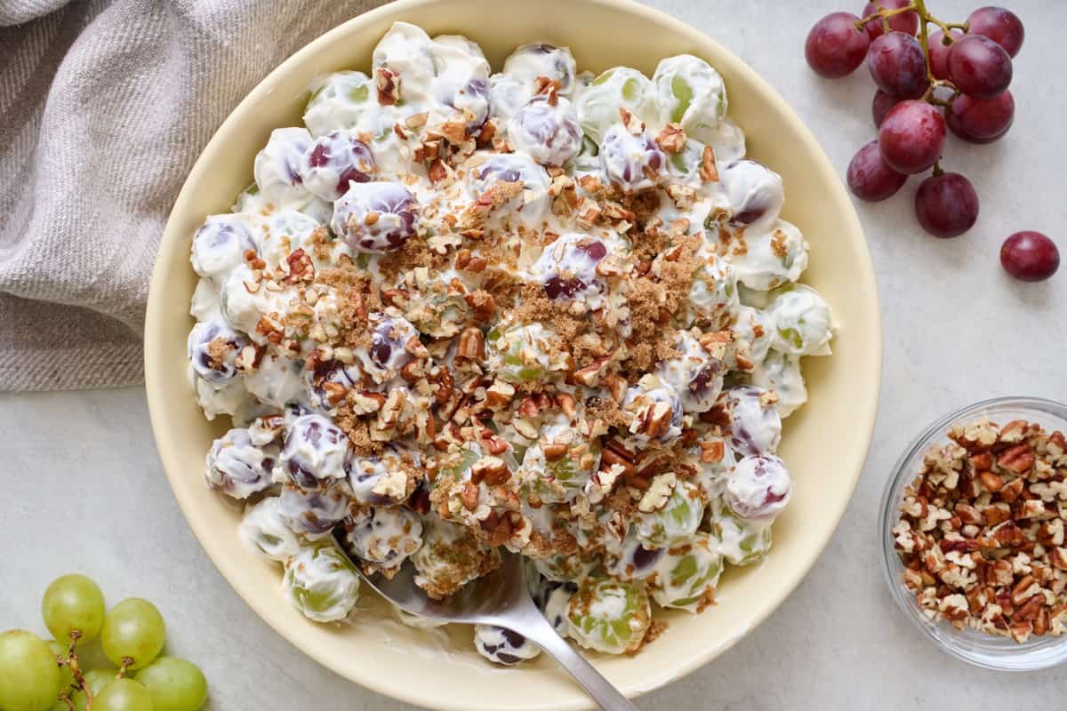 Grape salad piled up high in a small serving bowl topped with pecans and brown sugar with extra pecans and fresh grapes on the vine nearby.