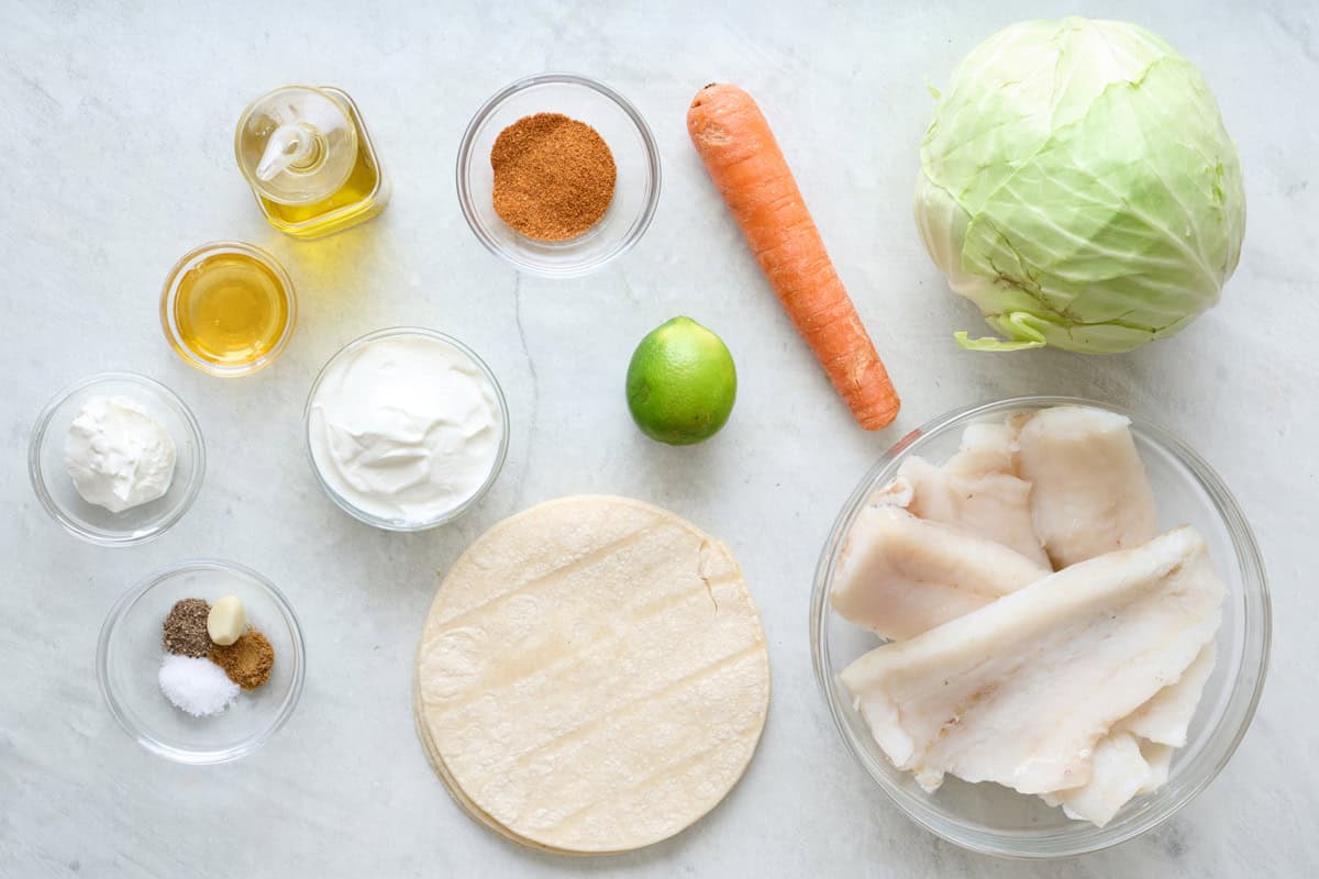 Fish taco ingredients on a marble countertop, including bowls of fish, taco seasoning, and oil.