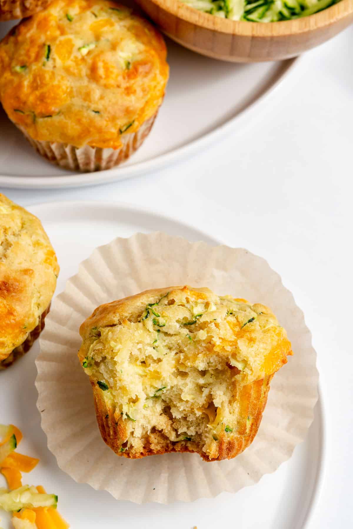 Zucchini and cheddar muffin on a small plate with a bite taken out and more muffins nearby.