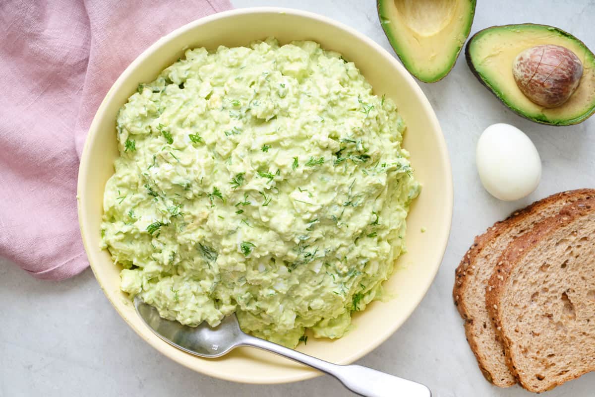 Avocado egg salad on a bowl with spoon dipped inside, fresh avocado, a boiled egg, and bread slices nearby.