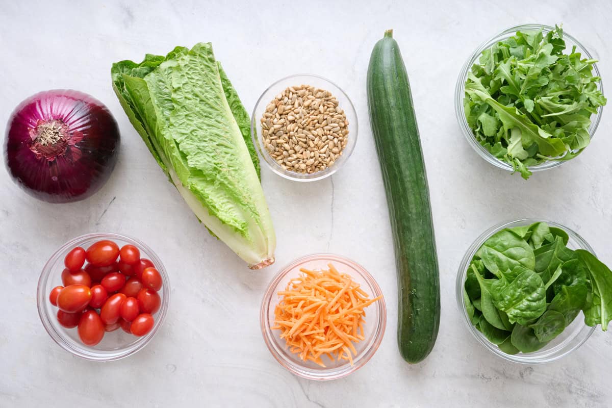 Ingredients for mixed greens salad: romaine lettuce, red onion, cherry tomatoes, grated carrot, cucumber, arugula, and spinach.