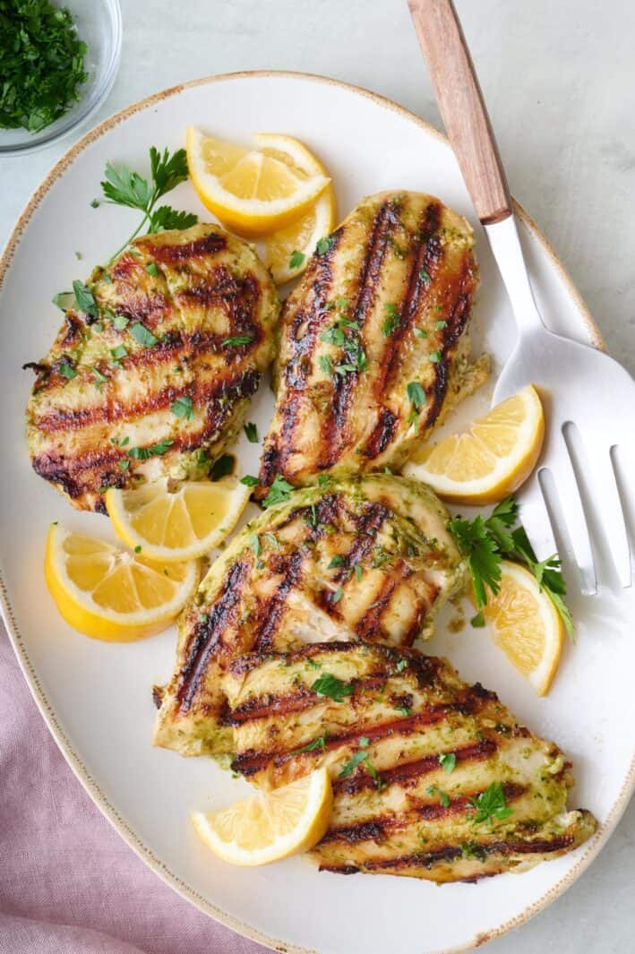 Pesto grilled chicken breasts on a platter, garnished with lemon wedges and fresh parsley.