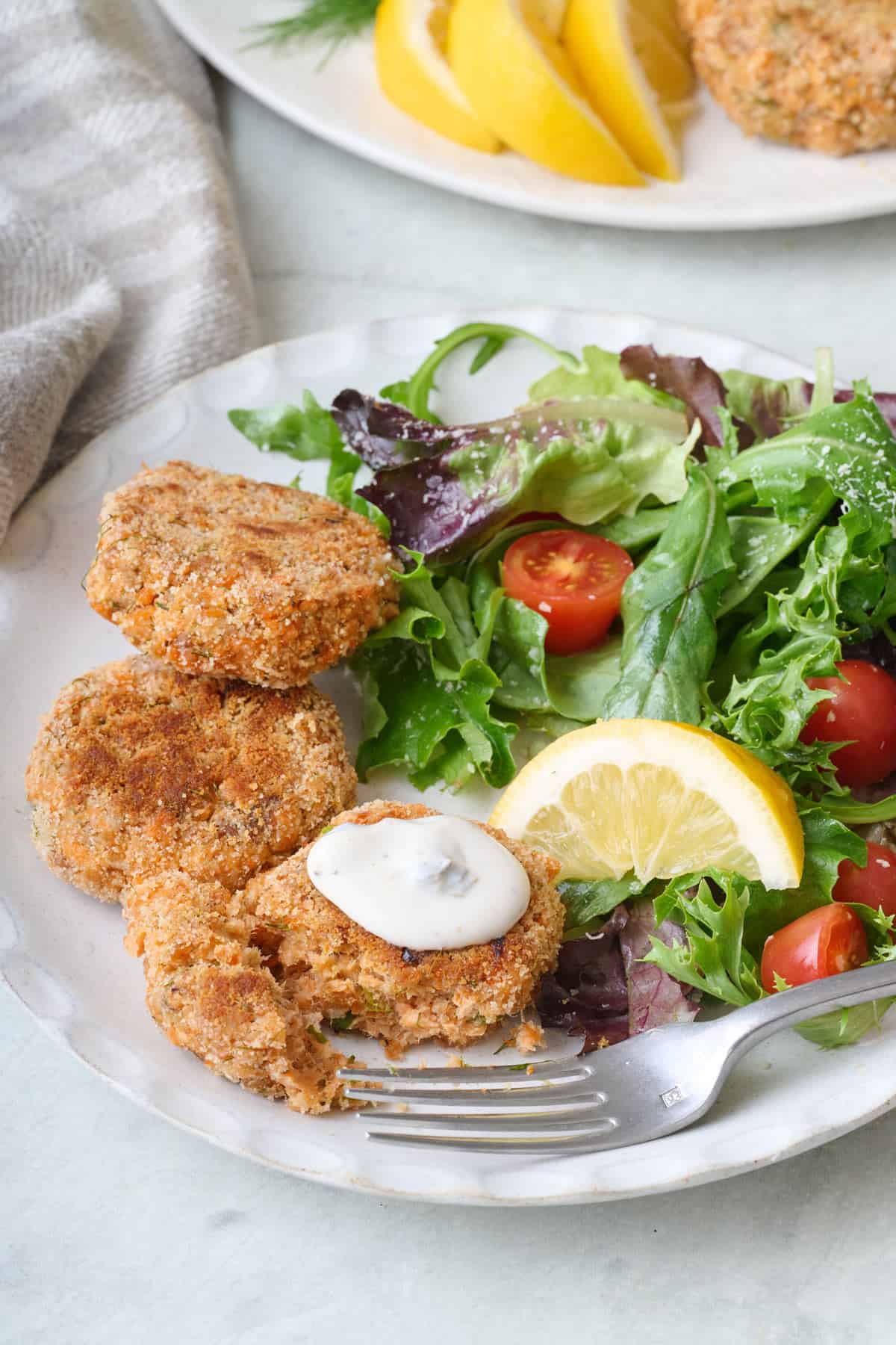A serving of pan-fried salmon patties on a plate with a bite flaked away to show inside texture and a dollop of yogurt sauce on top.