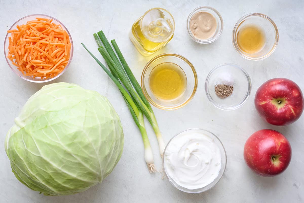 Ingredients for recipe before prepping: shredded carrots, cabbage, apples, green onions, oil, honey, apple cider vinegar, salt and pepper, and yogurt.