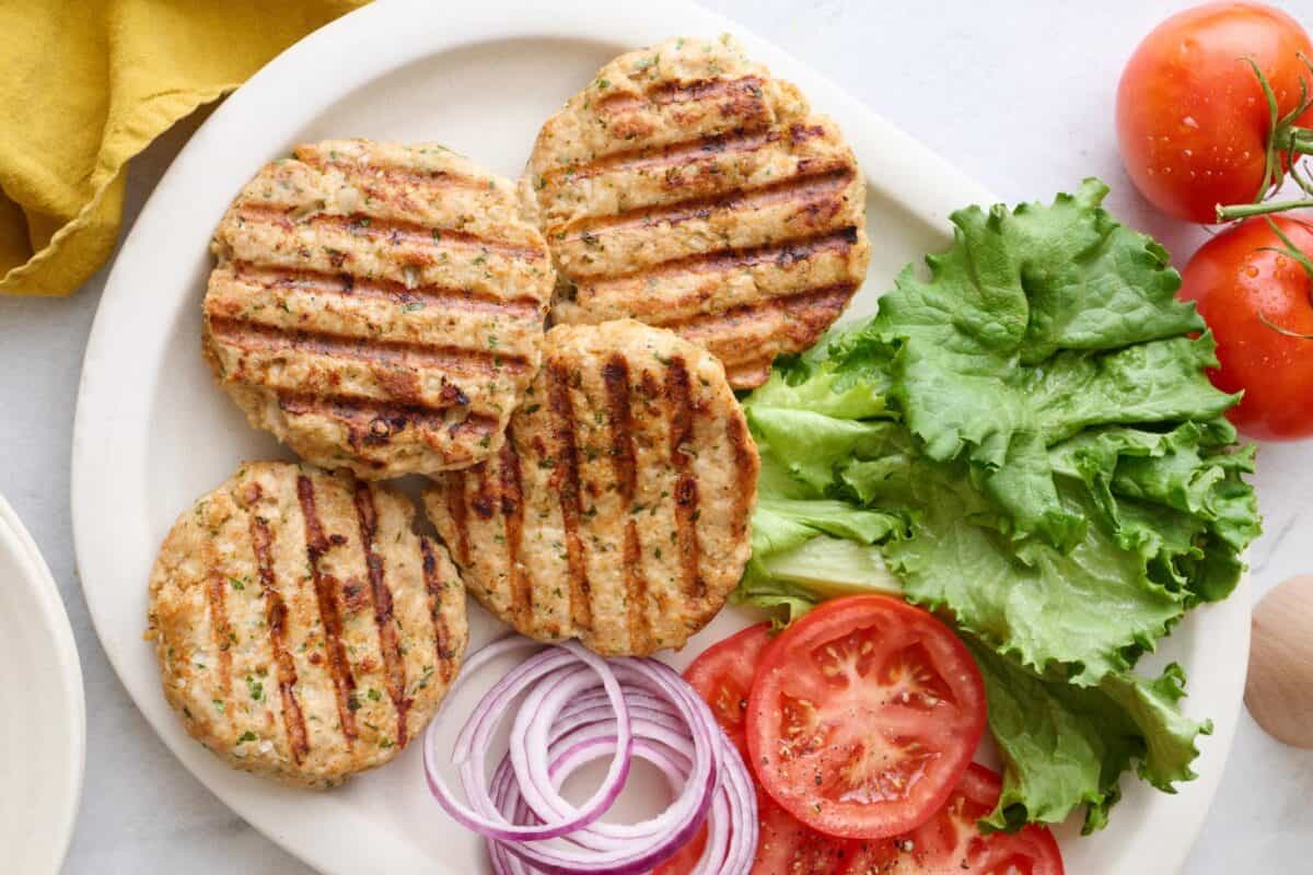Ground chicken burger patties on a platter with lettuce, sliced tomatoes, and sliced red onions for fourth of July.