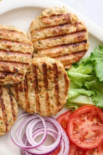 Ground chicken burger patties on a platter with lettuce, sliced tomatoes, and sliced red onions for fourth of July.