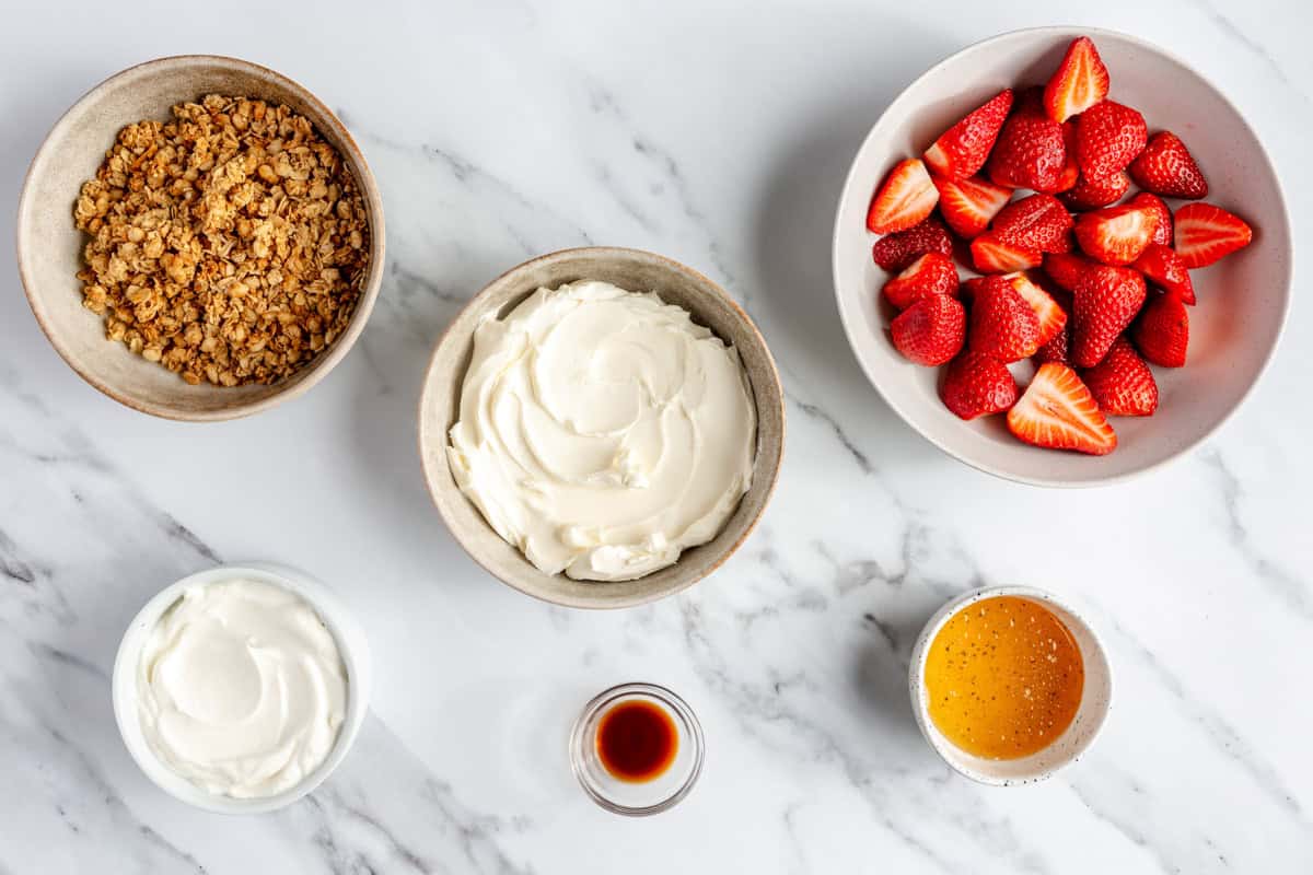 Strawberries, yogurt, cream cheese, honey, vanilla, and granola on a marble countertop.