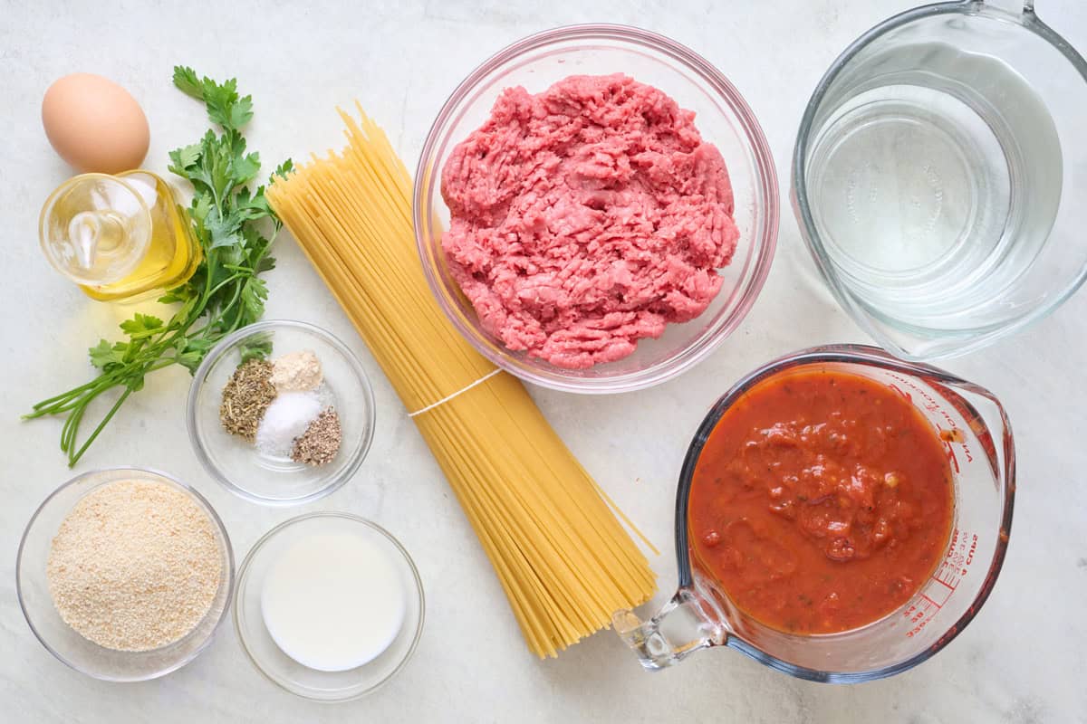 Spaghetti and meatball ingredients on a countertop top: pasta, marinara sauce, ground beef, breadcrumbs, herbs, spices, olive oil, egg, and water.