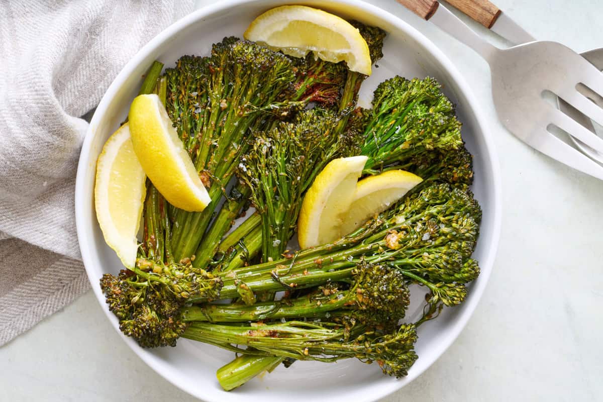 Bowl of seasoned roasted broccolini, garnished with lemon wedges.