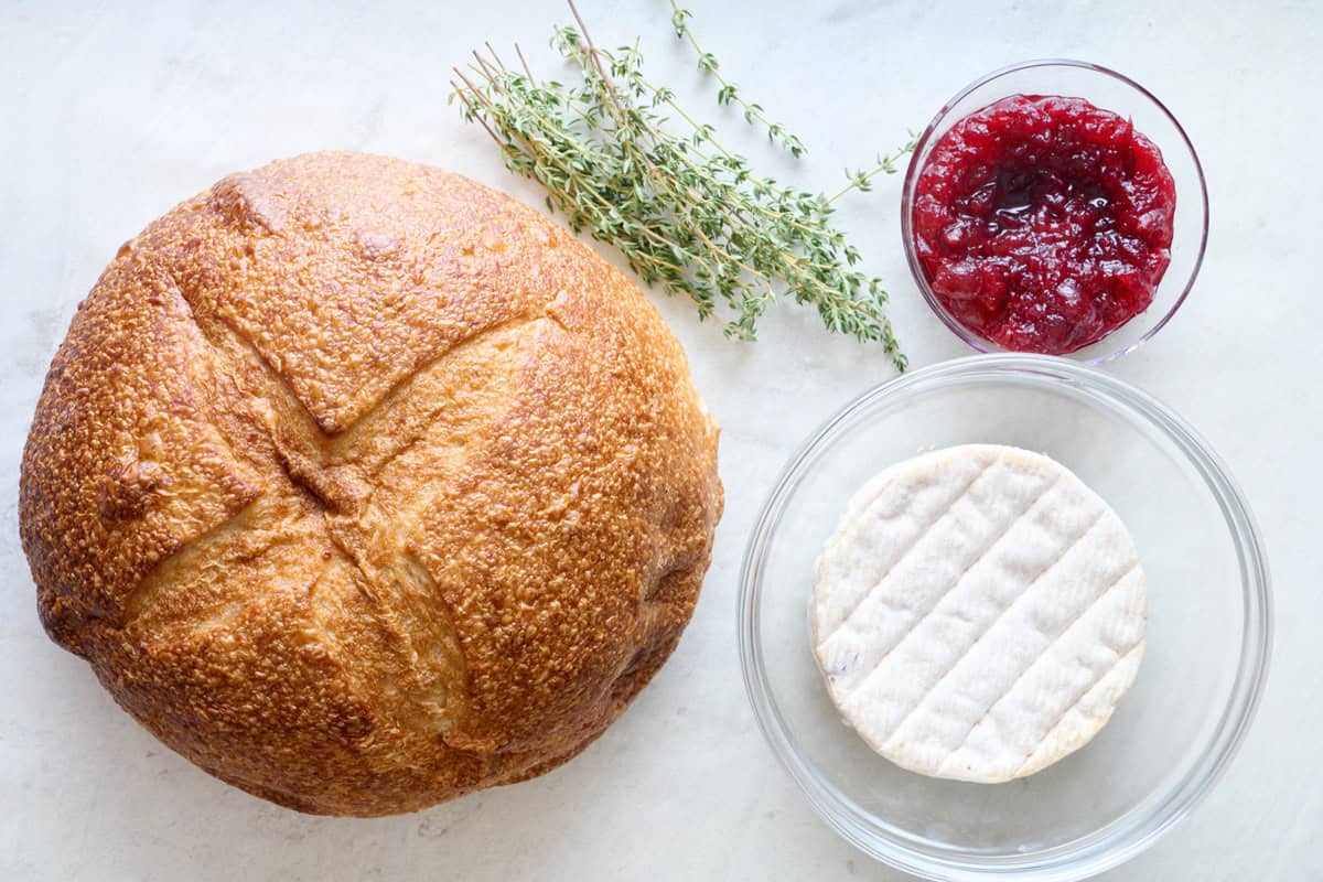 Ingredients for recipe: bread boule loaf, rosemary, cranberry sauce, and a brie wheel.