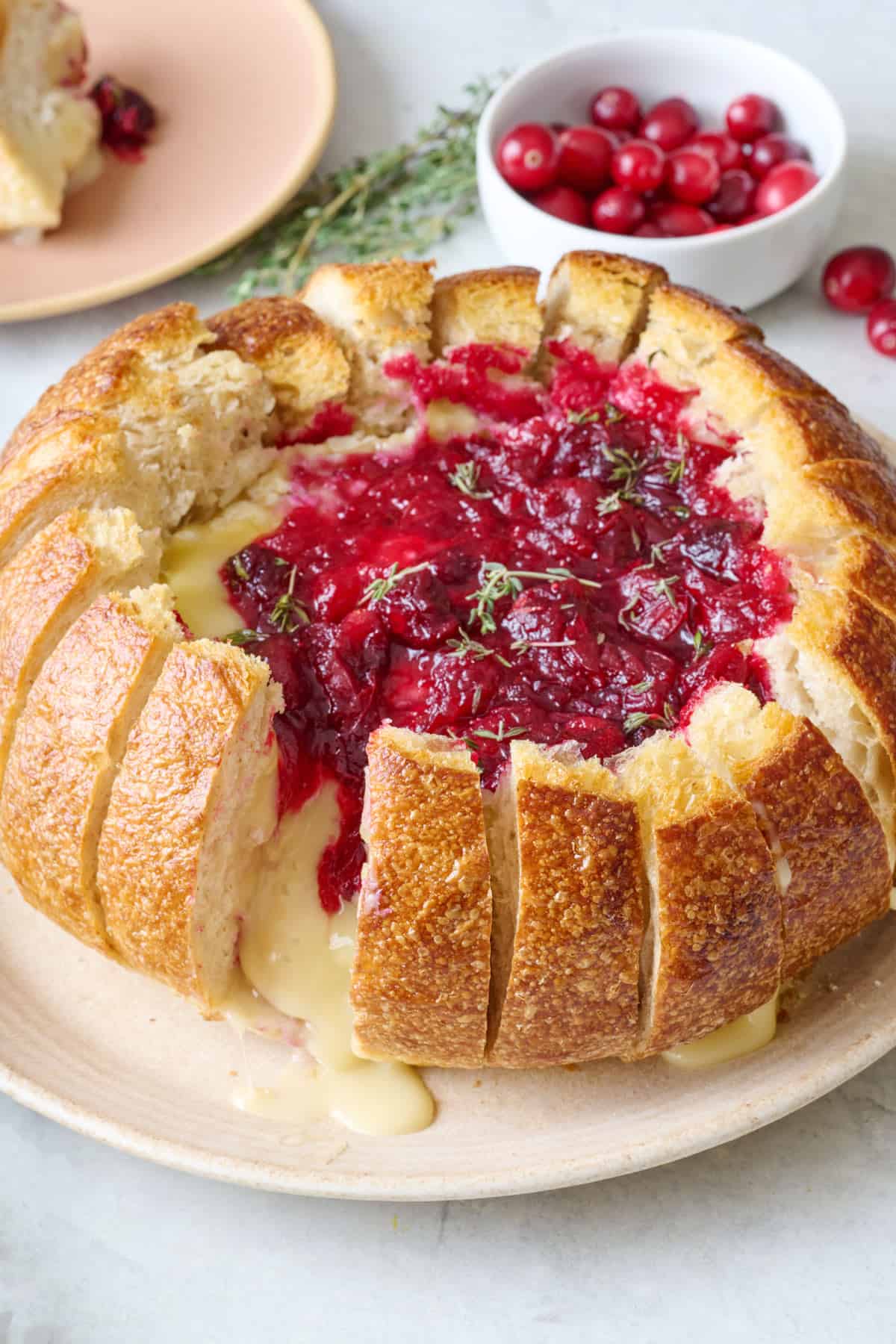 Baked brie in a bread bowl with cranberry sauce and rosemary and a few pieces of the sliced bread pulled away to show the melty cheese pouring out.