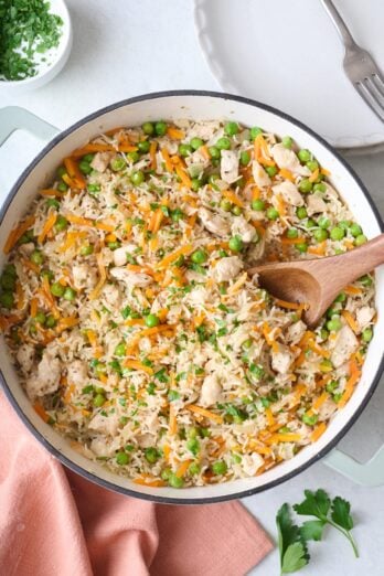 One pot chicken and rice in a white pot with peas, carrots, and herbs with a wooden spoon.