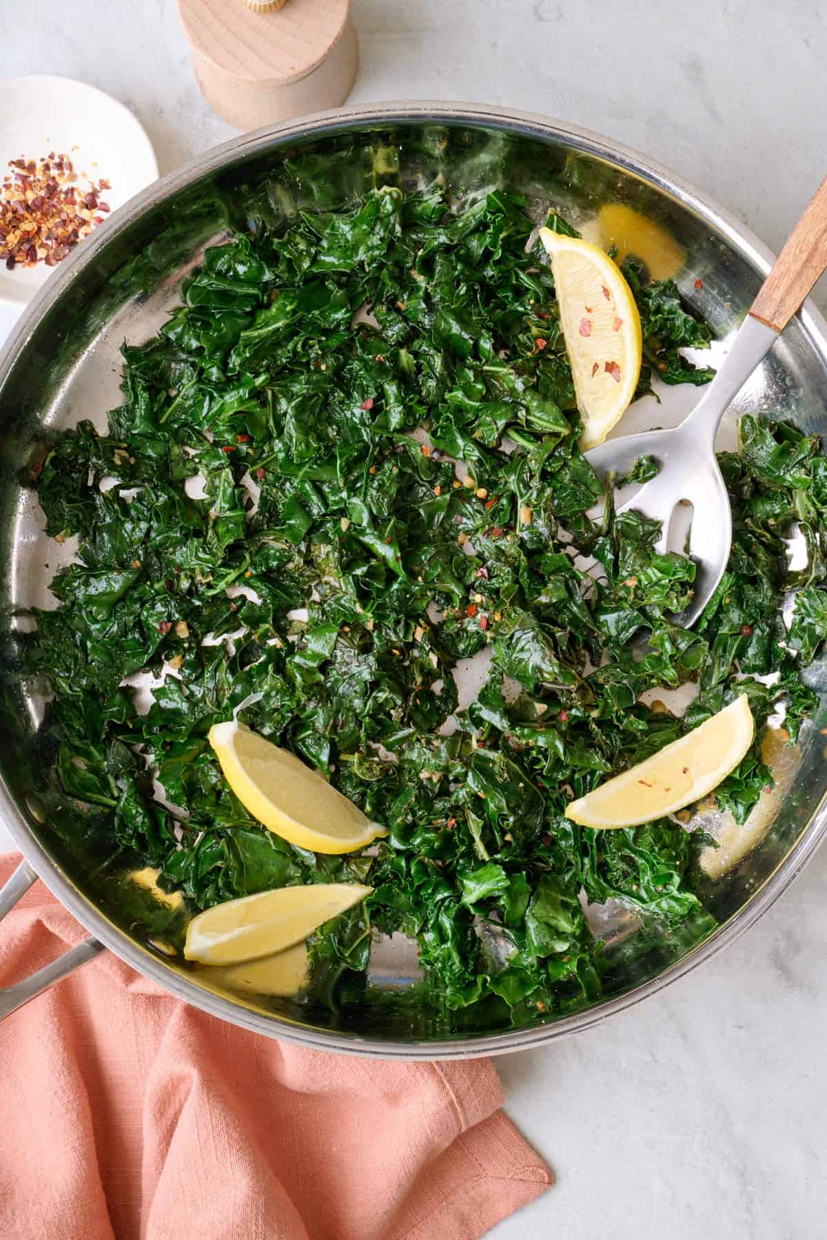 Sauteed kale in skillet with a few lemon wedges and serving spoon dipped inside pan.