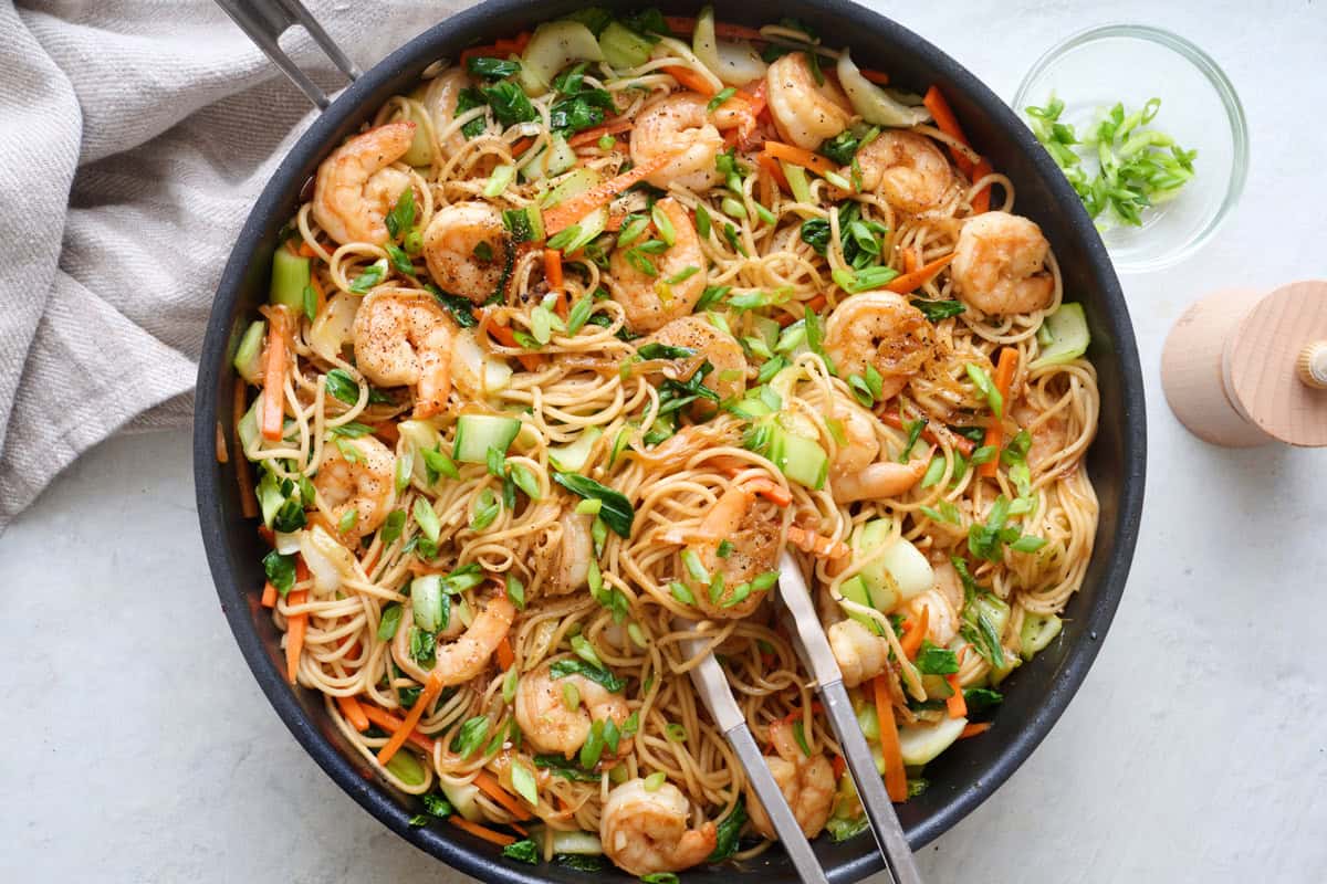 Shrimp lo mein in a skillet with tongs resting inside and more ingredients nearby.
