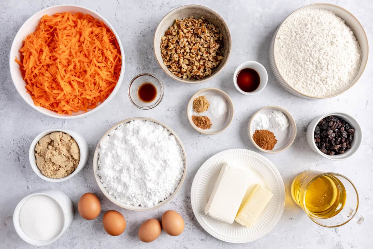 Ingredients for recipe in individual bowls: shredded carrots, brown sugar, sugar, eggs, powdered sugar, vanilla, walnuts, spices, cream cheese and butter, raisins, oil, and flour.