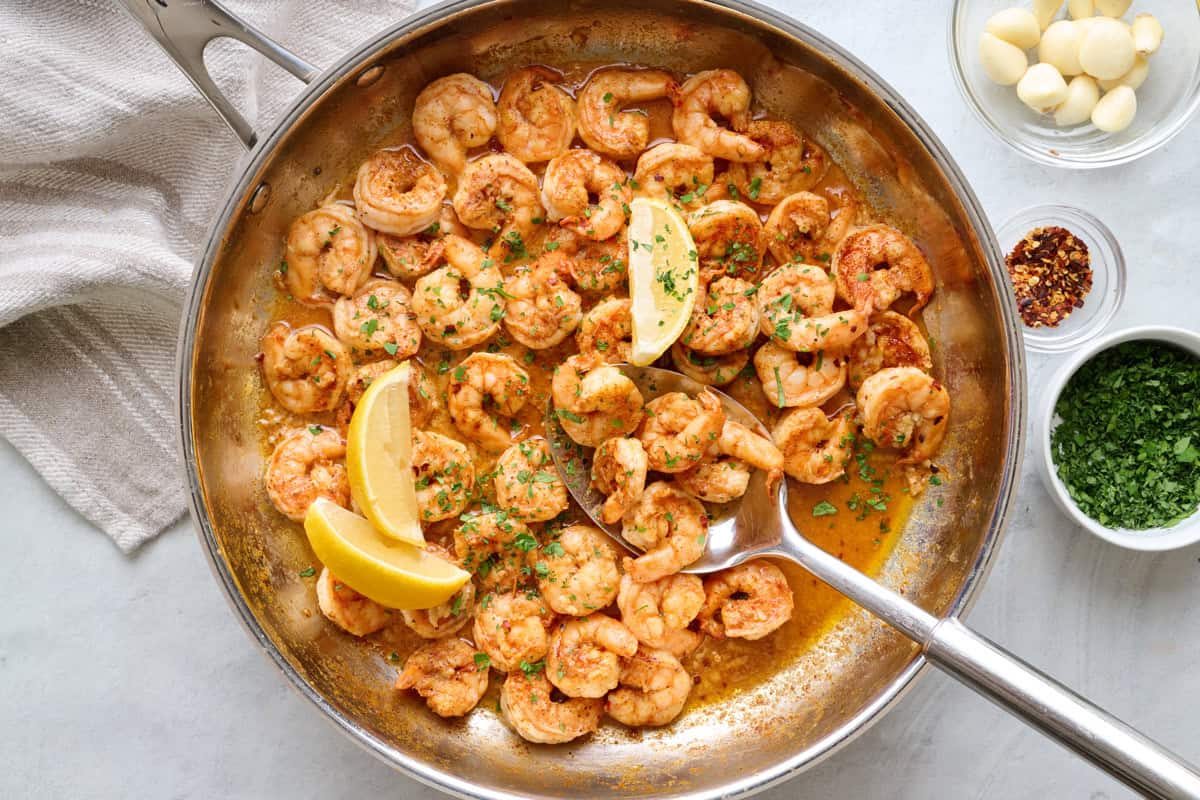 Shrimp in pan garnished with lemon wedges and chopped fresh parsley, garlic cloves nearby, large spoon dipped inside.