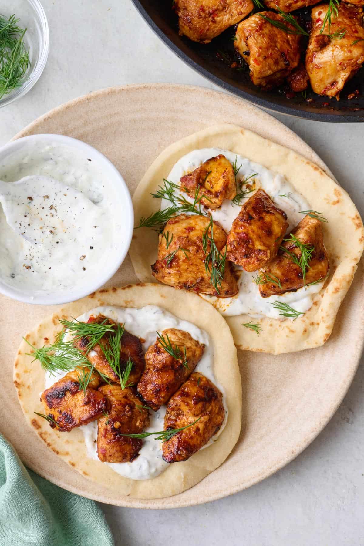 Two naans on a plate with harissa chicken pieces, yogurt sauce, and fresh dill.