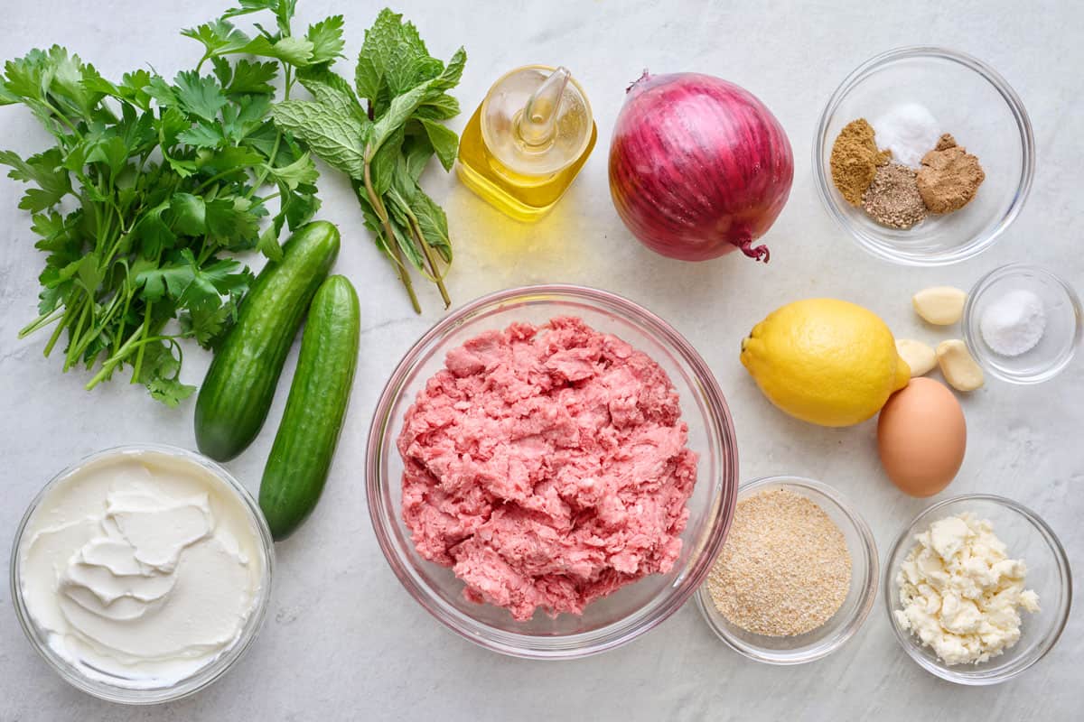 Ingredients for recipe: ground lamb, red onion, crumbled feta cheese, breadcrumbs, egg, garlic, fresh parsley, and spices with persian cucumbers, yogurt, garlic, lemon, oil, and mint for sauce.
