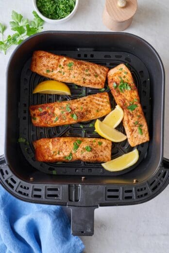 Air fryer salmon fillets in air fryer basket garnished with fresh parsley and lemon wedges.