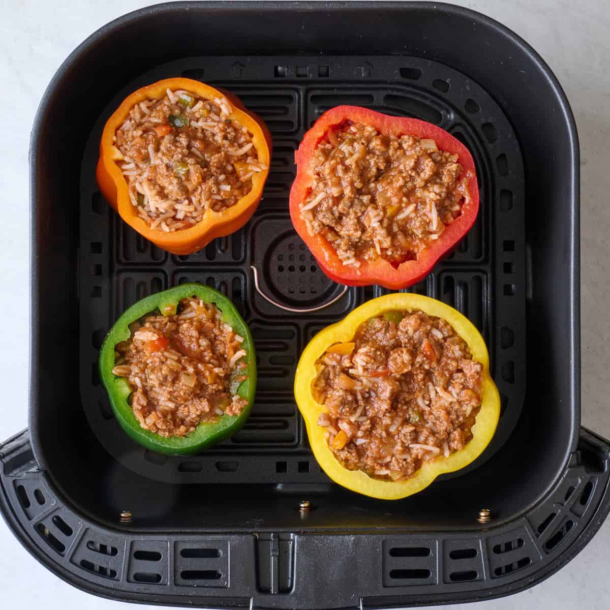 Four bell peppers stuffed with beef and rice filling before cooking in an air fryer basket.