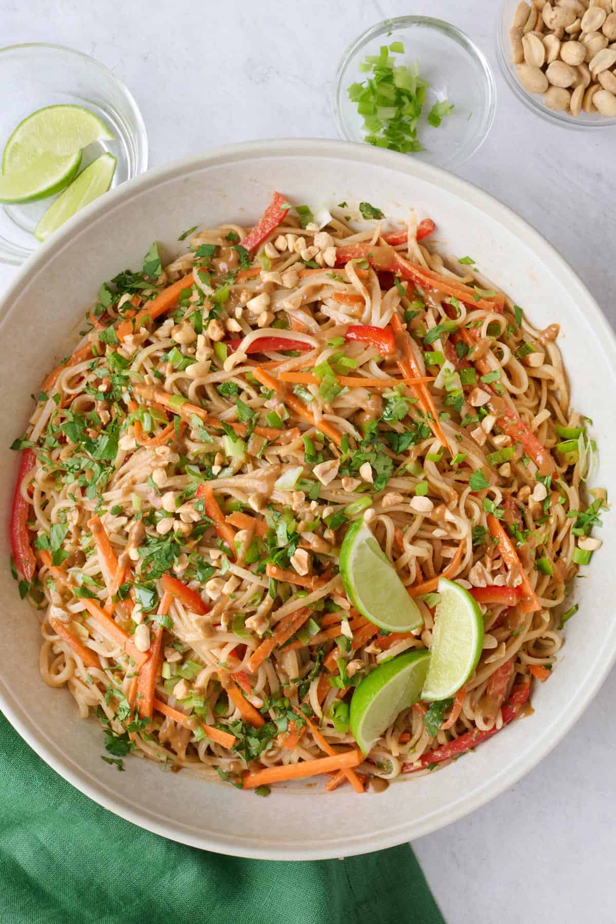 Asian noodle salad on a serving platter garnished with extra green onions, roasted peanuts and lime wedges, serving set tucked inside, jar of extra dressing and dish of peanuts nearby.