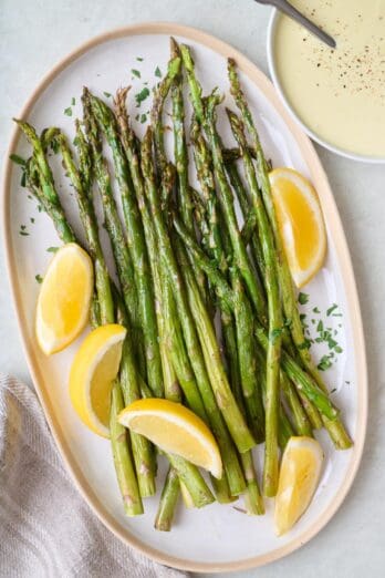 Roasted asparagus on a platter with lemon wedges and a bowl of hollandaise sauce nearby.