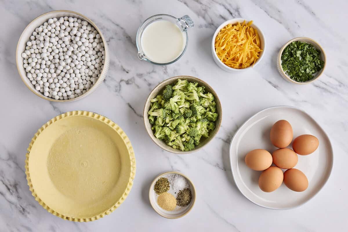 Ingredients for recipe: pie shell, pie weights, milk, broccoli, spices, shredded cheddar cheese, chopped parsley, and eggs.