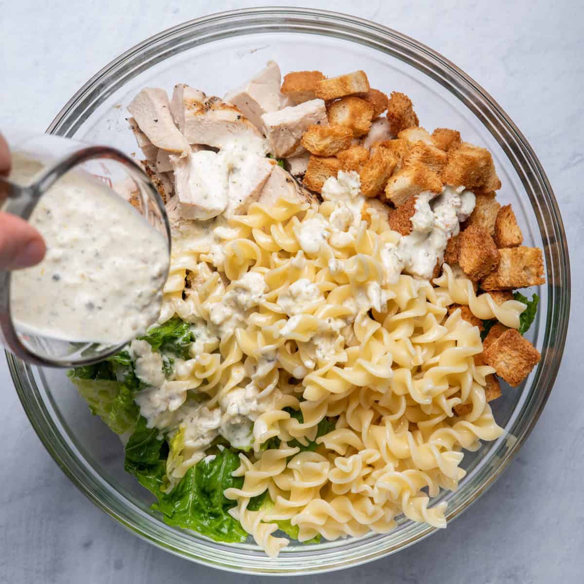 Bowl with compartments of lettuce, chicken, pasta and croutons and dressing being poured on top.