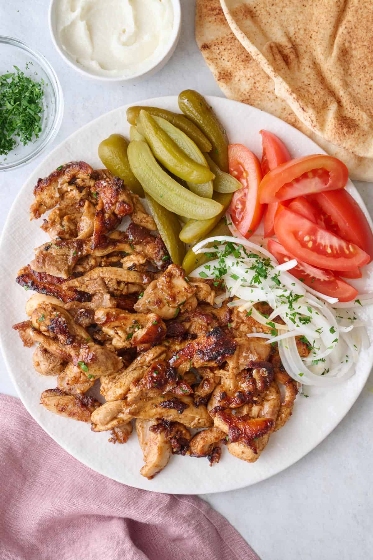 Chicken shawarma on a plate with pickles, tomatoes and onions with labneh and pita nearby.