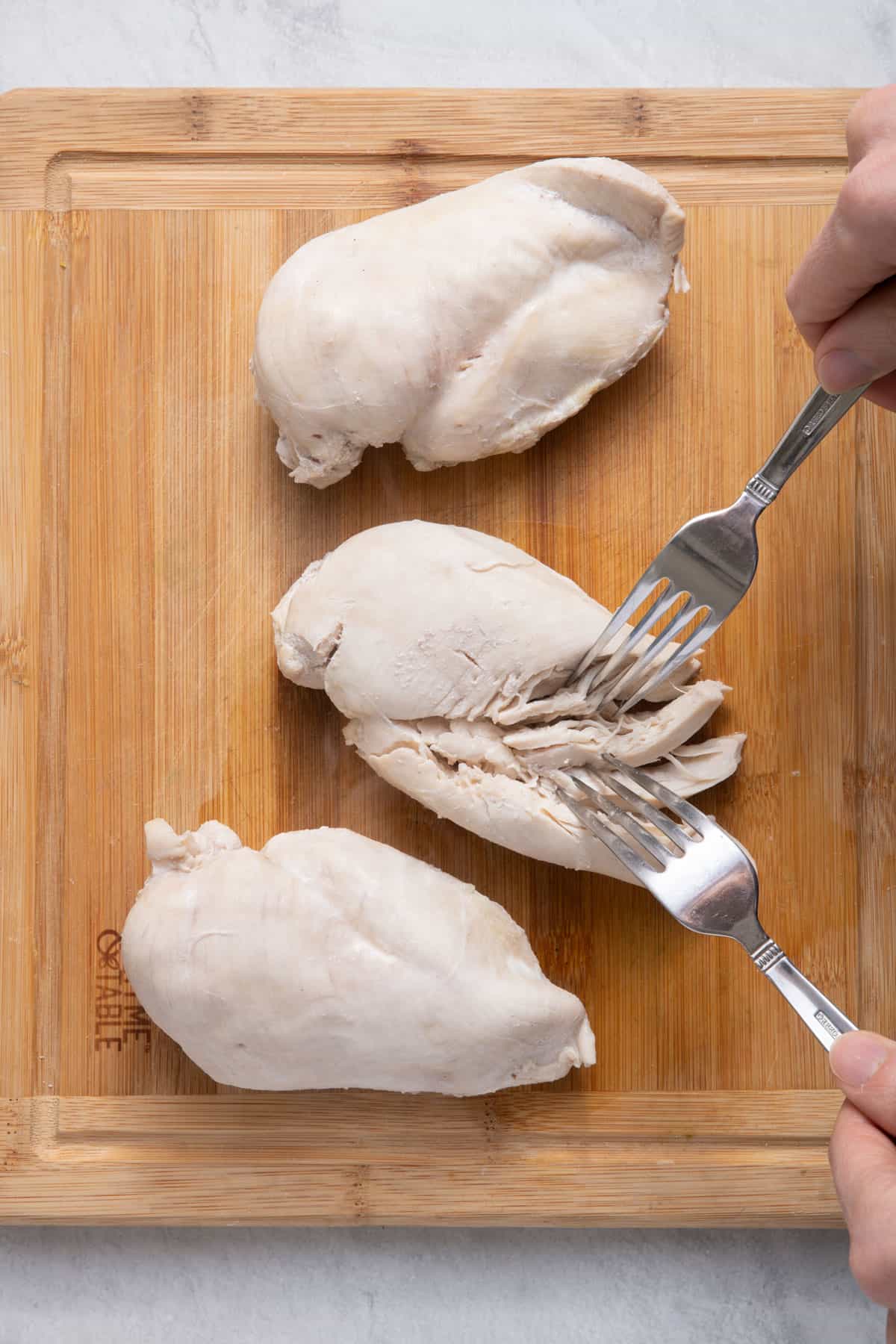 Two forks shredding a cooked chicken breasts on a cutting board.