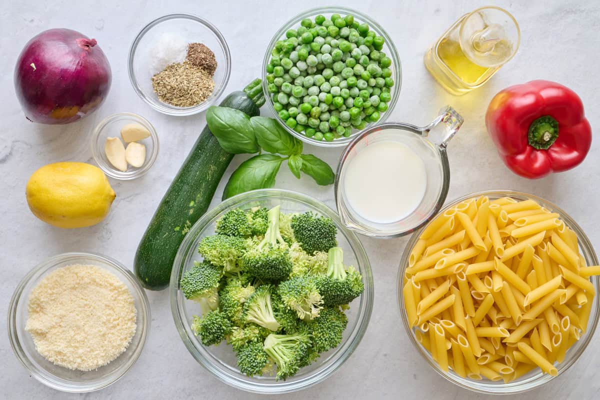 Ingredients for recipe: red onion, lemon, garlic, spices, parmesan cheese, zucchini, frozen peas, broccoli florets, milk, oil, red pepper, and penne pasta.