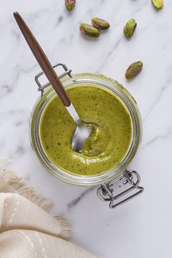 Pistachio butter in a small jar, spoon dipped inside, with extra shelled pistachios around.