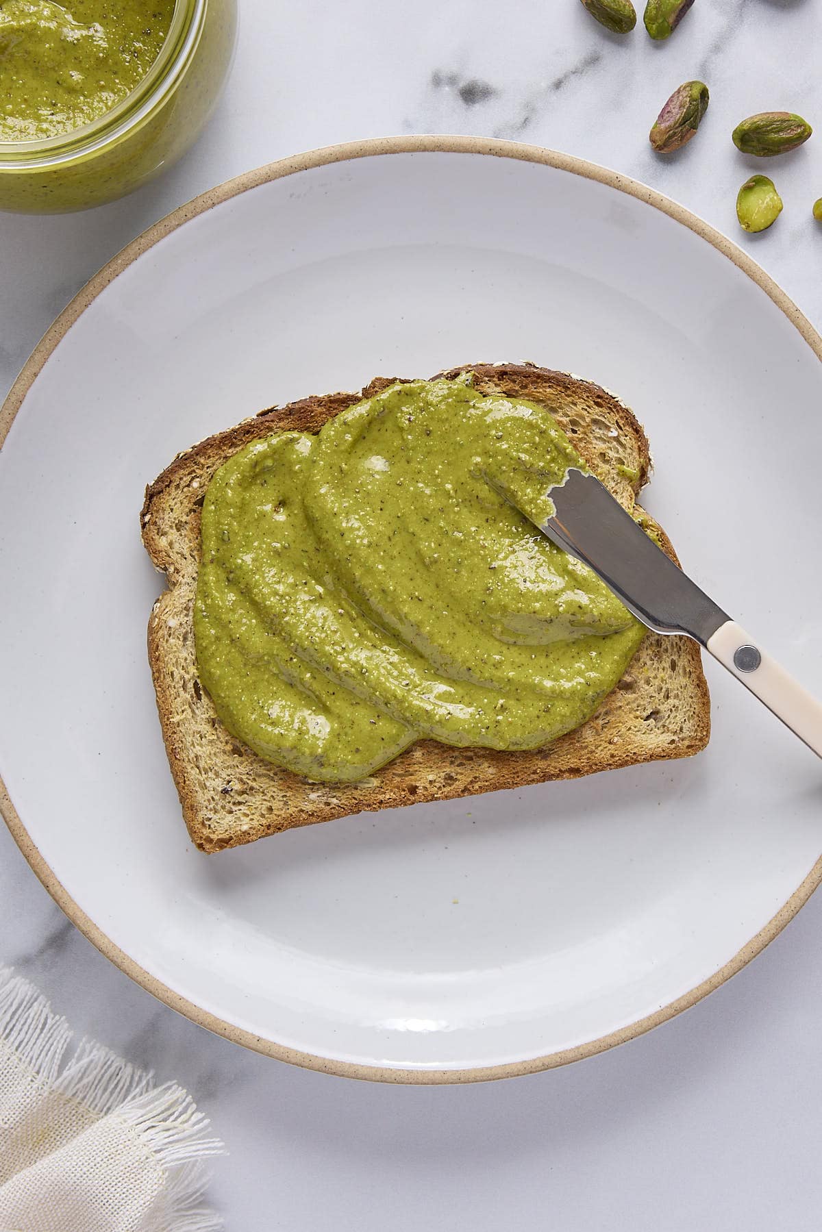 Spreading pistachio butter on hearty full slice of toast with jar nearby.