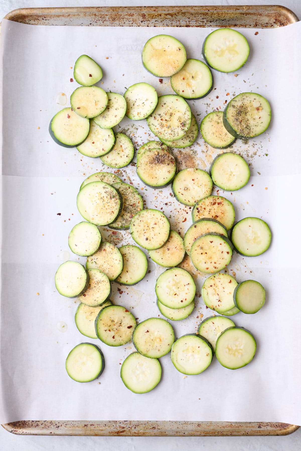 Sliced zucchini on a parchment lined sheet pan with oil and seasonings added before tossing together.