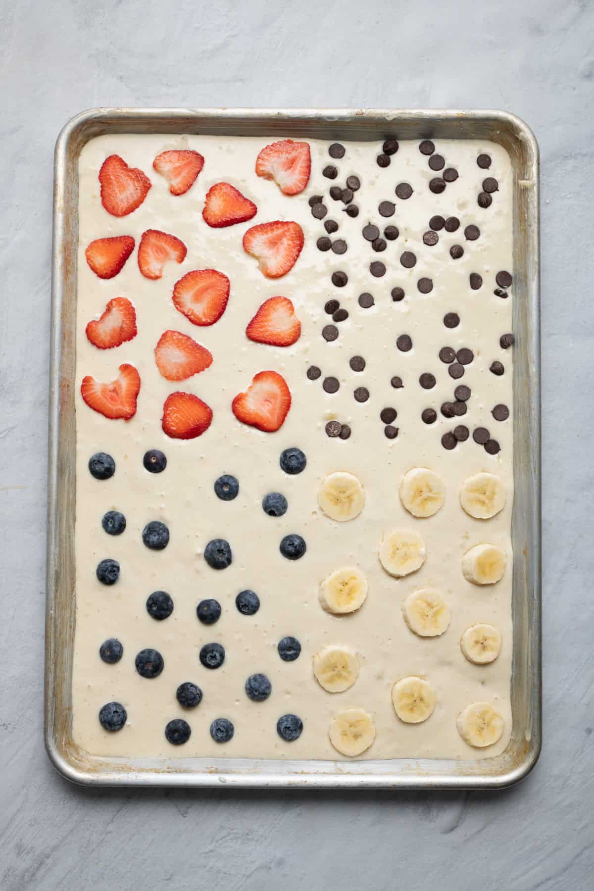 Pancake batter on a sheet pan with different toppings in each section: strawberries, chocolate chips, blueberries, and bananas.