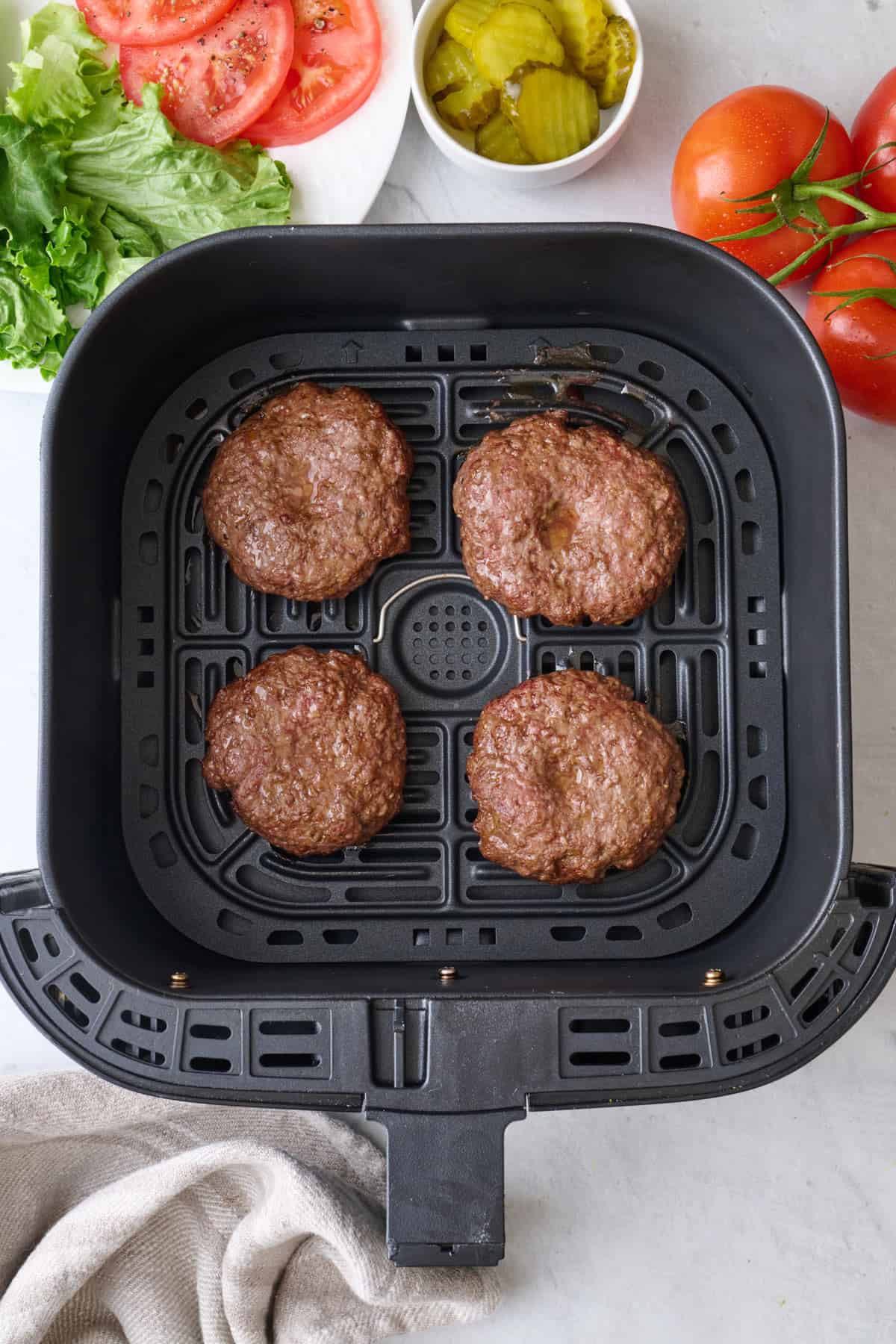 Hamburger patties in an air fryer basket with burger toppings nearby.